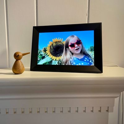 A black picture frame sitting on a mantle, displaying a photo of a girl wearing sunglasses standing next to a sunflower.
