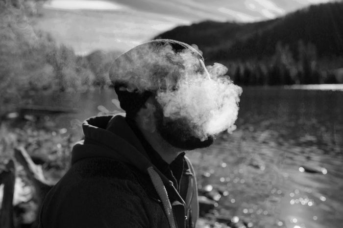 Person in a beanie and hooded jacket exhaling smoke or vapor by a lake in a serene outdoor setting.