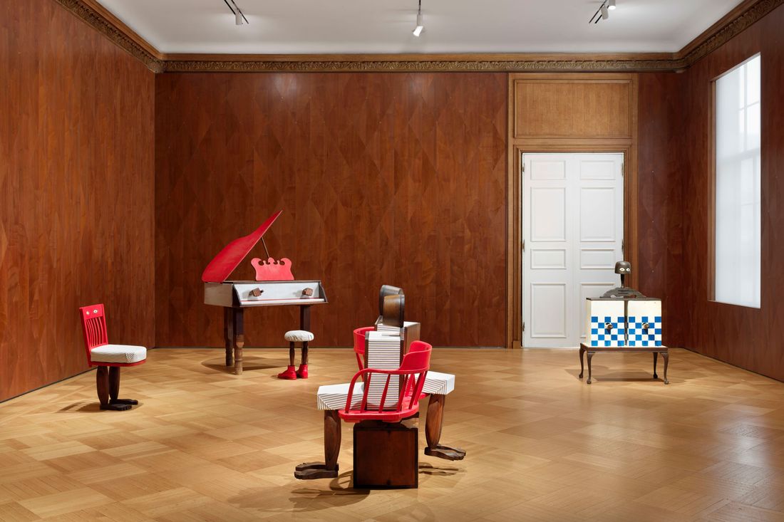 Furniture-like sculptures are in a gallery with wood-paneled walls and a wood floor. The works include a cabinet with a blue-and-white checkerboard front, a Victorian-style love seat in which to wooden sculptures of people face opposite directions, a non-playable piano, and a stool with legs that look like legs in boots.