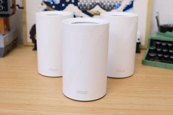 3 TP Link Deco router nodes sitting on a wooden desktop, arranged in a triangle.