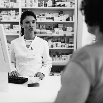 In this photo, a pharmacist stands at a computer behind a counter. She is mid-speech and a box of medication rests on the counter in front of her. A patient stands with their back to the camera in the foreground.