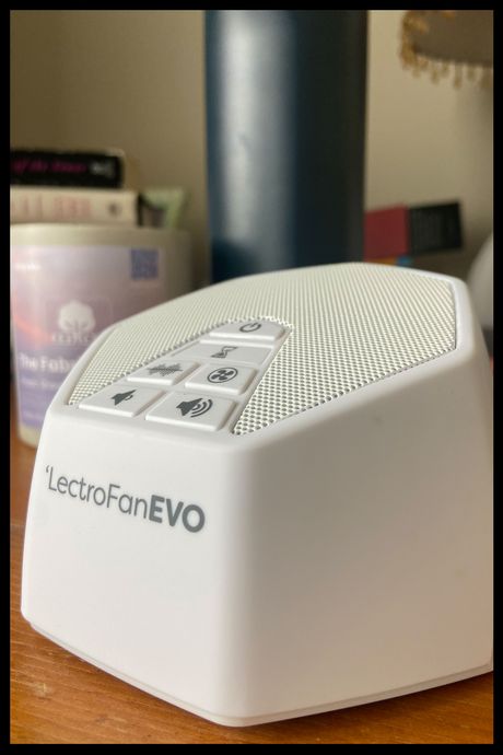 A white sound machine on a nightstand with a candle and books in the background.