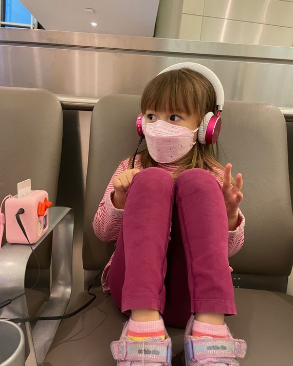 Un niño con auriculares y una máscara facial se sienta en un asiento del aeropuerto escuchando el audio de un reproductor de audio Yoto Mini.