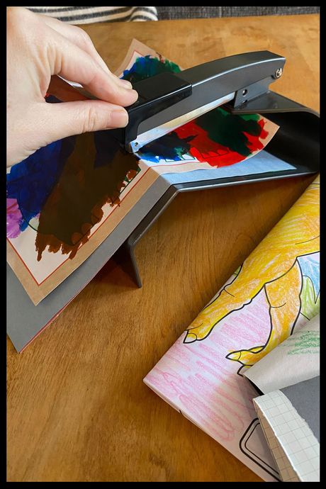 Photo of a woman's hand using a booklet stapler to make zines out of toddler artwork.