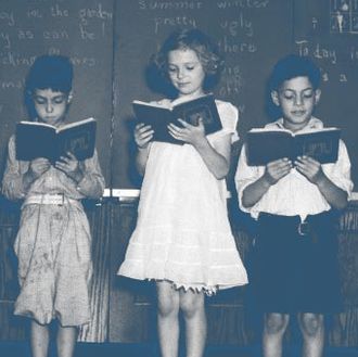 1930s line-up of 5 elementary school students in front of blackboard reading books with teacher looking on