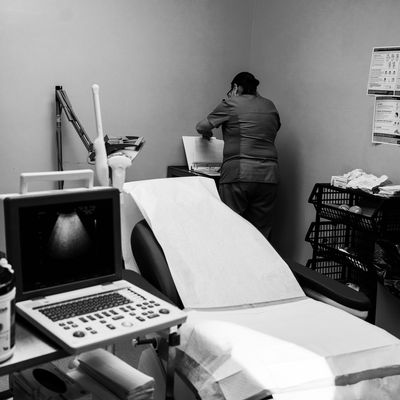 An empty exam chair in an abortion clinic. A worker in the background, who has her back turned, flips through the pages of a binder.