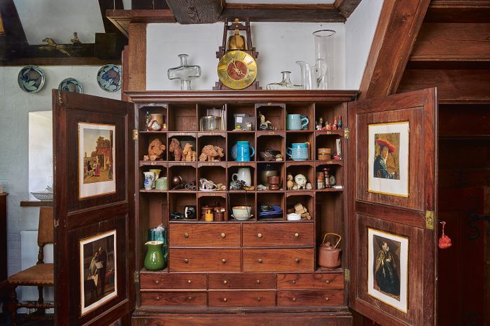 A wooden cupboard open with small enamelware, carved wood, and ceramic cups on its shelves with closed drawers below.