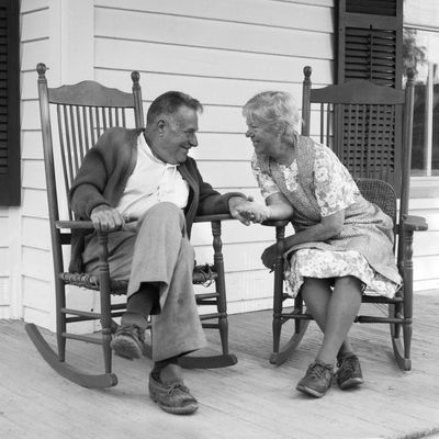 1970s elderly couple in rocking chairs on porch holding hands