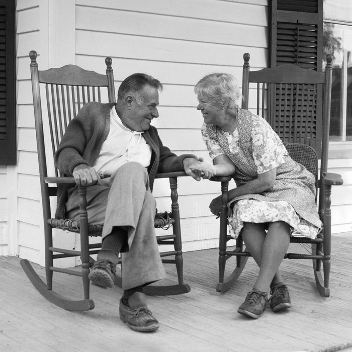 1970s elderly couple in rocking chairs on porch holding hands