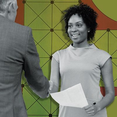 A professionally dressed woman holds a piece of paper in her left hand as she shakes hands with a man standing in front of her.