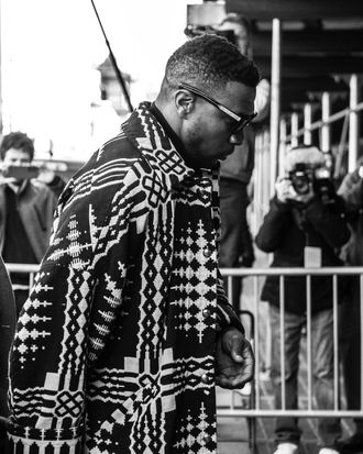 A black man wearing sunglasses and a long, patterned coat walks in front of a barricade. Photographers behind the barricade snap his photo.