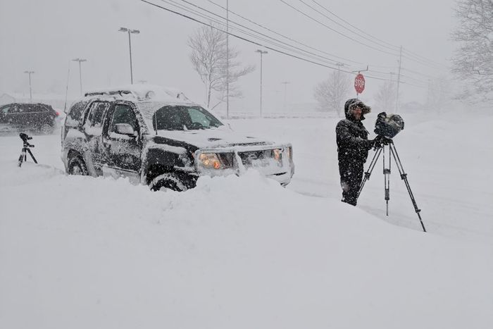 Man with tripod and camera standing in front of car in deep snow.
