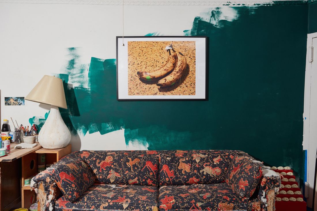 A room with a partially painted teal-green wall, a red-and-black paisley print sofa, a photograph of a ripe banana hanging on the wall, and the corner of a desk with jars of paint brushes and a table lamp with a tilted shade.