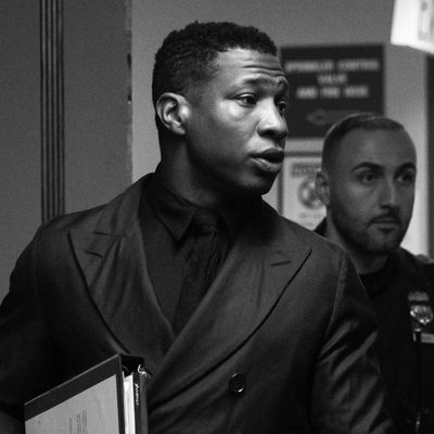 Actor Jonathan Majors, wearing a black collared shirt and a black overcoat, walks into a courtroom holding a binder. 