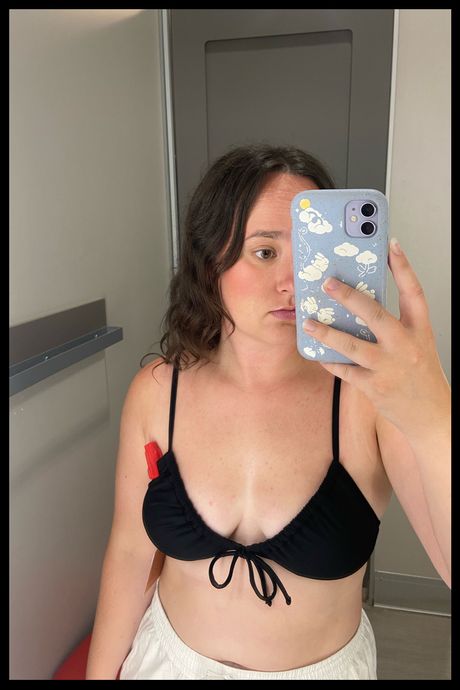 A person trying on a black swimsuit top in a dressing room.
