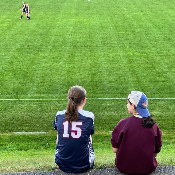 Two girls sit on a path at the edge of a soccer field.