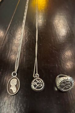 Una imagen de primer plano de un collar con la silueta de una persona, otro collar y un anillo, cada uno de los cuales tiene la misma L cursiva grabada.