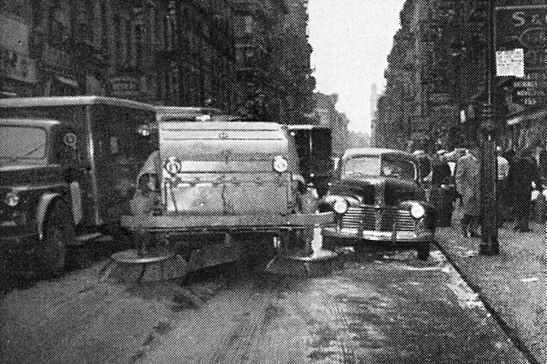 A black and white image of a street sweeping vehicle trying to squeeze between two parked cars 