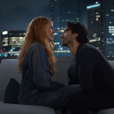 A woman and a man sit on a rooftop with a city skyline spreading out behind them. They are looking into each other's eyes, and the man is leaning in toward the woman's face.