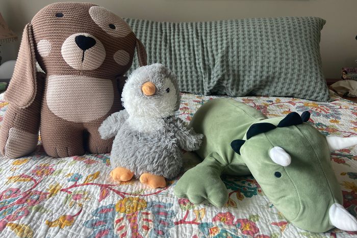 From left to right, the Darcy the Dog plush, Gray Penguin Warmies plush, and Pillowfort Green dinosaur plush in a row on top of a bed..