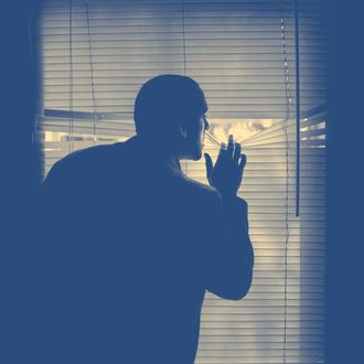 A man peeking outdoors through the blinds of a darkened room