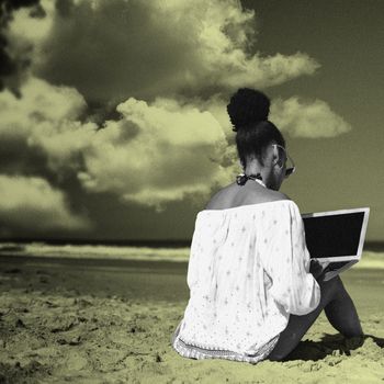 A woman sits on the beach and types on a laptop balanced on her knees. Her hair is in a bun and she's wearing an off-the-shoulder coverup over her bathing suit and sunglasses. She faces the waves and her back is to the camera. 