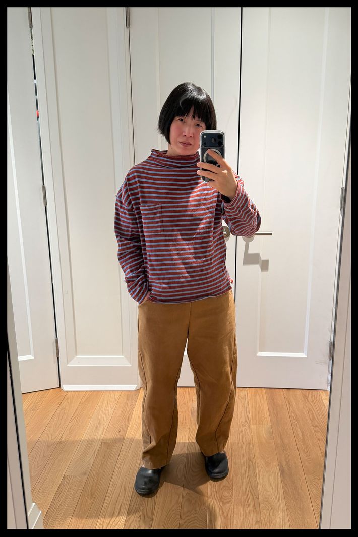 A woman with short dark hair taking a mirror selfie while wearing a red and blue striped sweater and a pair of Le Bonne Shoppe Arc Pants in the tobacco color.