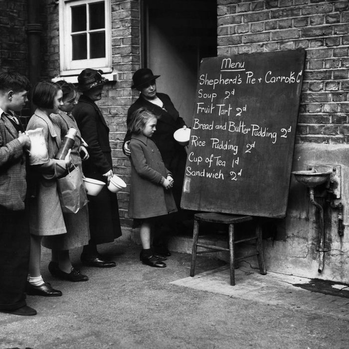 A vintage black-and-white photograph of women and children in a line, looking at a short food menu written on a chalkboard