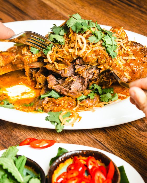 A lamb shank bathed in an orange-colored curry sauce, falling off the bone and sprinkled with fried shallots, cilantro, next to small bowls of herbs and peppers.