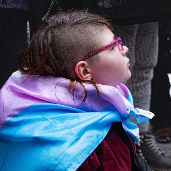 A young girl with half her hair buzzed poses for a portrait from the side, wrapped in a trans pride flag.