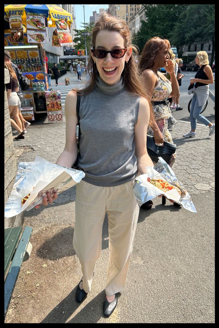 A woman at a street fair holding wrapped hotdogs in each hand while wearing a gray sleeveless top, sunglasses, flats, and a pair of cream Brooke Callahan Tie Pants.