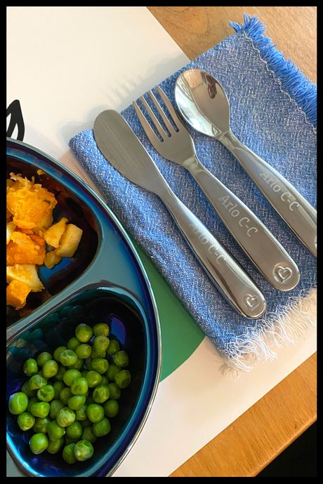 Photo of a set of three stainless steel toddler utensils — fork, spoon, knife — on a napkin next to a plate of peas and sweet potatoes.