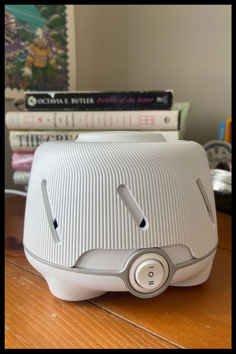 A white sound machine on a nightstand with books in the background.