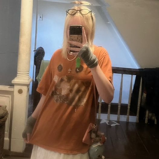 A teenager poses for a portrait in the mirror, wearing an oversize orange T-shirt and covering their face with their phone.