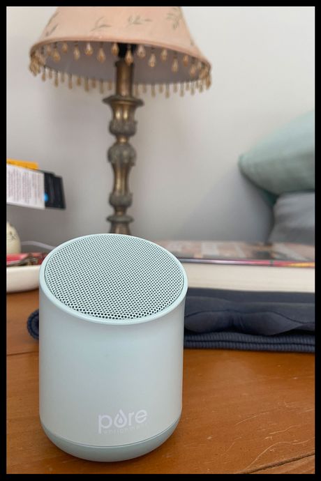 A green sound machine sitting on a bedside table, with a lamp and pillow in the background.
