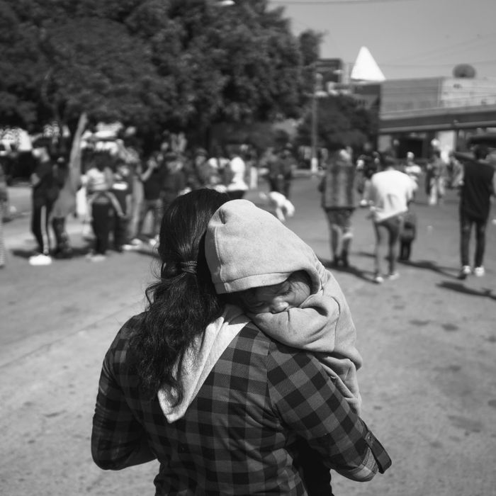 A woman wearing a plaid hoodie walks with a child resting on her right shoulder at the El Chaparral border crossing in Tijuana, Mexico.