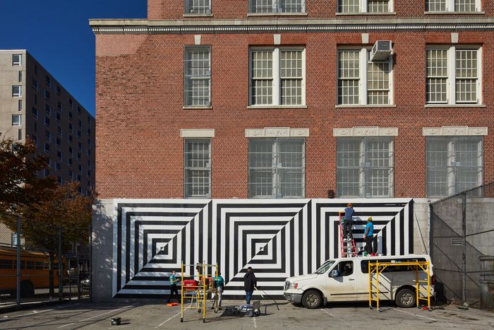 A dozen students in blue shirts installing a black-and-white optical-art mural on a brick building while using a cherry picker