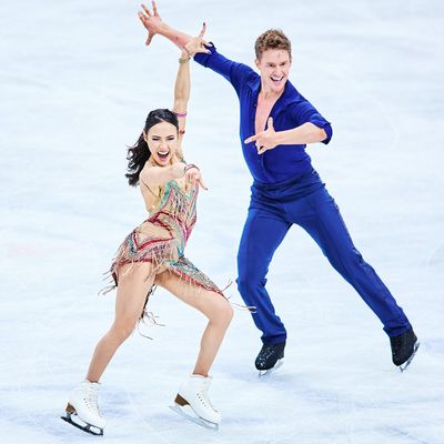 A woman and a man are ice dancing in a rink. The woman has her brown hair pulled into a ponytail and is wearing a nude costume covered in multi-colored beaded fringe. The man is wearing a blue velvet jumpsuit with a plunging v-neck. 