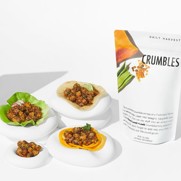 Two packages of Daily Harvest crumbles are pictured. Walnut + Thyme and Leek + Lentil crumbles are laid out in four small bowls, on top of pasta, a lettuce wrap, and a tortilla.