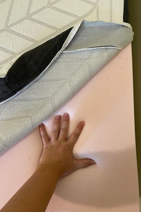 A mattress topper with the cover unzipped to show the foam, and a person's hand pressing into it.