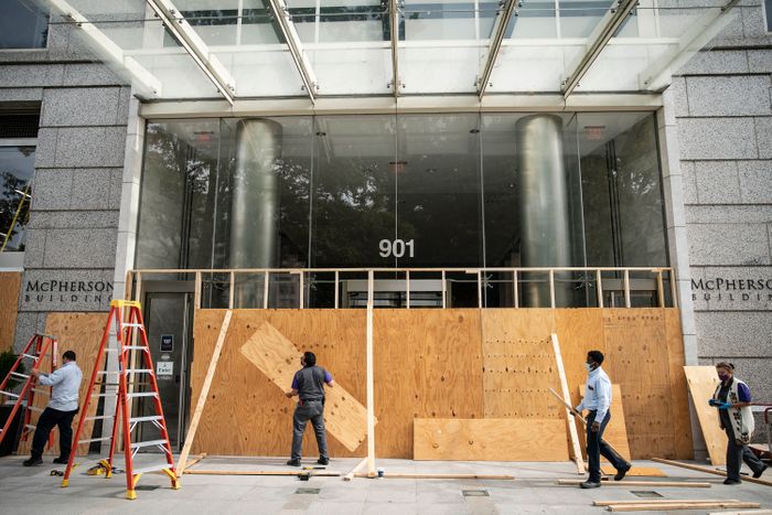 Workers in front of a large commercial building are placing plywood in front of windows.