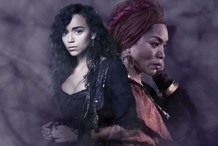 ID: against a cloudy background on a dark screen, two people who read as women stand with their backs against each other, one facing forward and one facing to the side. Their hair and skin are dark. The one facing forward has a black corset on and short, dangly earrings. The one facing to the side has a gold bangle on her hand that reaches to her head, is wearing longer hoops and an orange top, and she is wearing a head wrap.