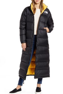 A full length The North Face Nuptse Long Water Repellent Down Coat with a bright yellow lining and The North Face logo over a cream colored sweater, blue jeans, and navy slip on sneakers on a model. The Strategist - 48 Things on Sale You’ll Actually Want to Buy: From Sunday Riley to Patagonia