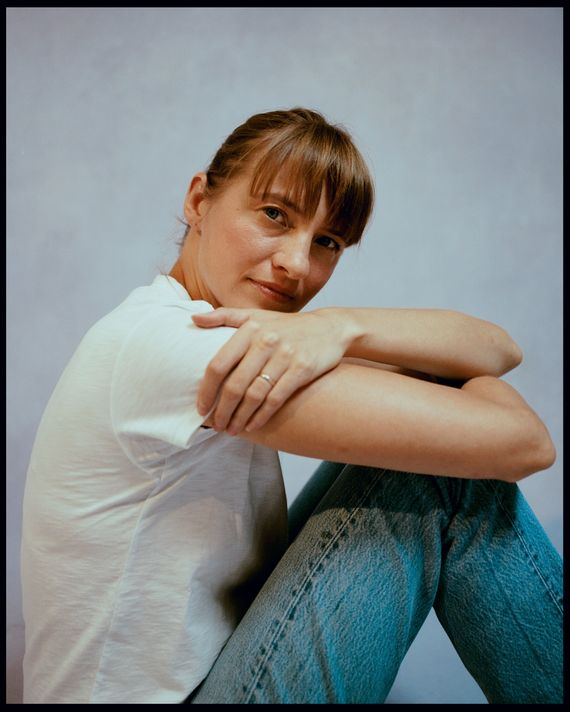 A woman wearing a white tee shirt and jeans poses for a portrait. Her brunette hair is pulled back and she has bangs covering her forehead. She is sitting on the ground and resting her arms on her knees, turning her head to the right to look at the camera.