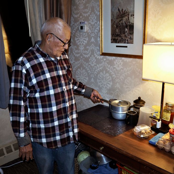 An elderly man with white hair looking at the top of a dresser with a pot, lamp, and packaged food.