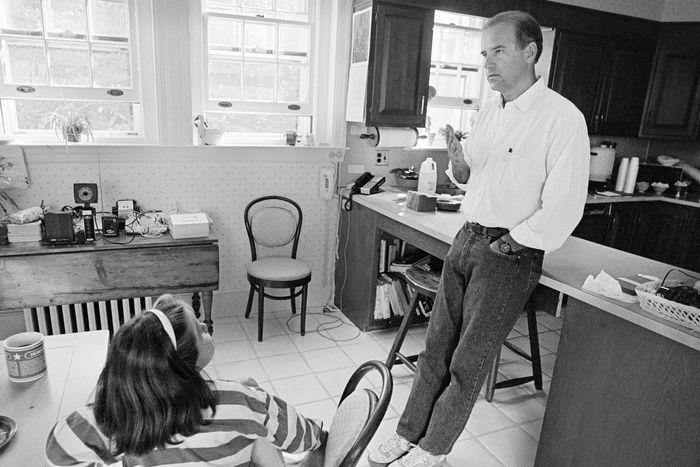 Joe Biden leans against a counter in his kitchen. His young daughter is sitting down at a wood kitchen table talking to him.