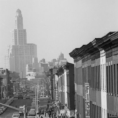 View of the street with people milling about and traffic at the intersection of Claver Place and Fulton Avenue in the Bedford Stuyvesant section of Brooklyn, 1968. The Williamsburgh Savings Bank tower can be seen in the distance.