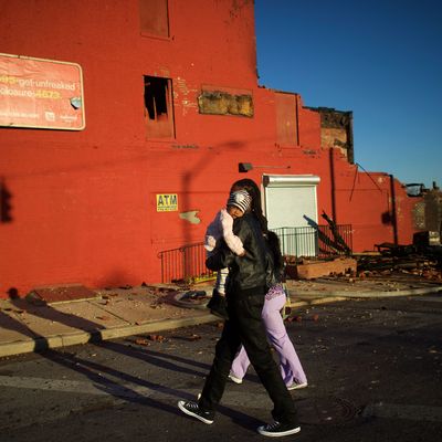 BALTIMORE, MD - APRIL 28: A man carries a young child past the remains of convenience store and a senior center set ablaze during night riots smolder at dawn on April 28, 2015 in Baltimore, Maryland. A state of emergency has been declared in Baltimore following violent protests which erupted after the funeral service for Freddie Gray. Gray, 25, was arrested for possessing a switch blade knife on April 12 outside the Gilmor Homes housing project on Baltimore's west side and according to his attorney died a week later in the hospital from a severe spinal cord injury he received while in police custody. (Photo by Mark Makela/Getty Images)