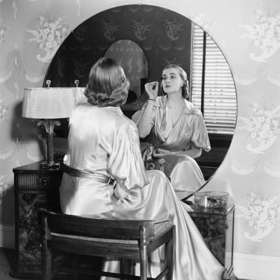 Woman applying make up in front of vanity table, (B&W),