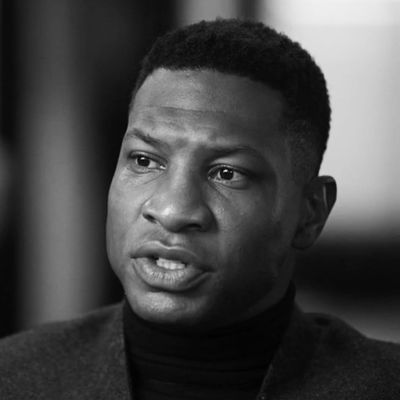 A black man wearing a turtleneck and suit jacket looks tensely off-camera.